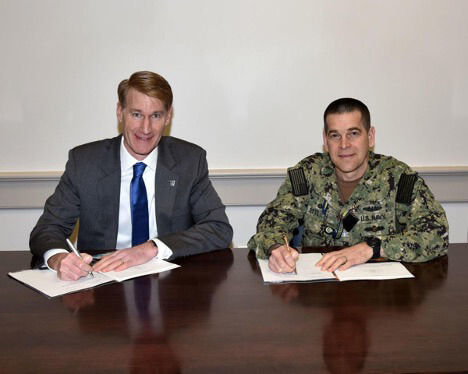 Two men sitting at a wooden desk preparingto sign a document while posing