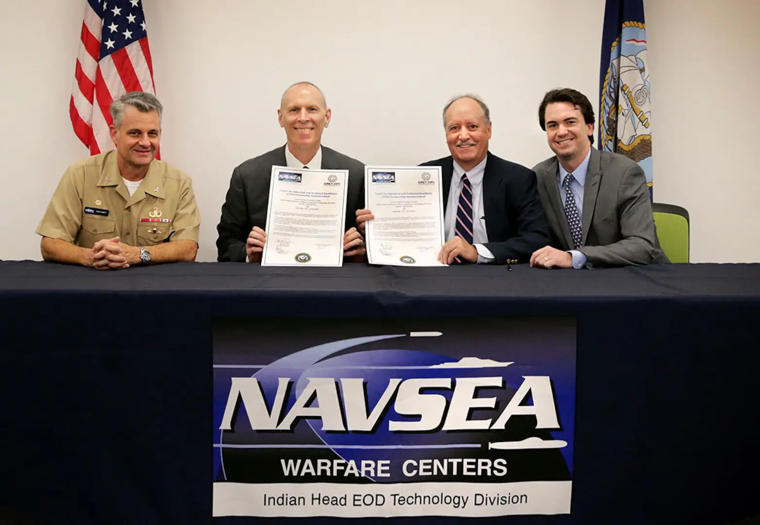 Four men sitting at a table with a banner hanging down that reads, "NAVSEA Warfare Centers. Indian Head EOD Technology Division"