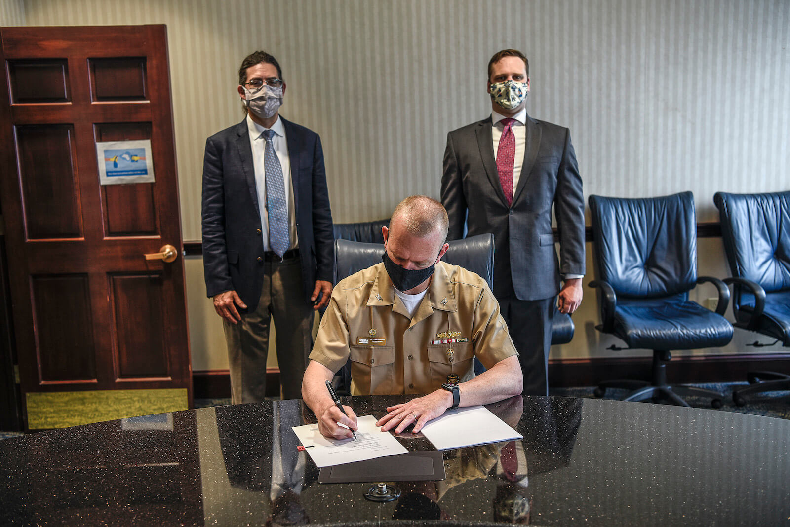 A man sitting at a wooden desk signing a document, while two men stand behind him in medical masks posing